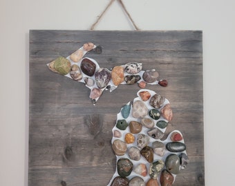 May include: A wooden sign with a white outline of the state of Michigan. The outline is filled with various colored stones. The sign is labeled "Maggie Shay 2023".