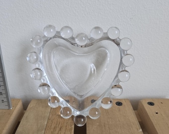 May include: Clear glass heart-shaped dish with a beaded edge. The dish is sitting on a wooden surface. The heart-shaped bowl is approximately 4 inches wide.