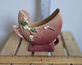 May include: A vintage, pink ceramic vase with a unique crescent shape. The vase features a decorative floral design with white berries and green leaves. It has a small handle and sits on a rectangular base. The vase is displayed on a wooden surface.