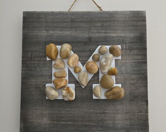 May include: A wooden sign with the letter "M" made from small, light brown stones. The sign is hanging from a rope and has a gray wood background.