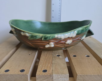 May include: A green and brown ceramic bowl with a leaf and floral design. The bowl has a boat-like shape with a dark green interior and a decorative rim. The exterior features a textured, brown surface with white and green accents.