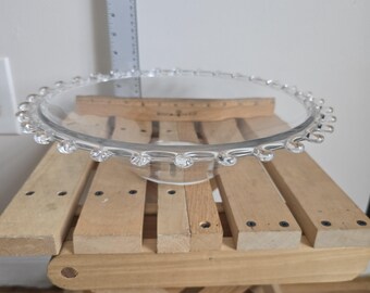 May include: A clear glass cake stand with a decorative beaded edge. The stand is round and sits on a small base. A wooden ruler is placed on the stand. The stand is on a small wooden folding table.