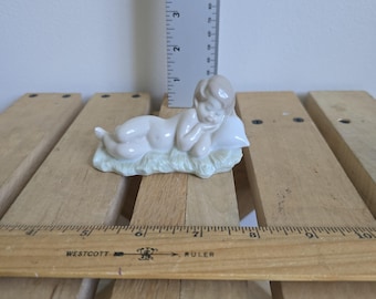 May include: A porcelain figurine of a reclining cherub, resting on a white pillow and a bed of light green grass. The cherub is nude, with a light brown hair and a serene expression. The figurine is approximately 3 inches long.