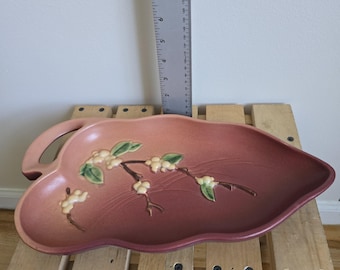May include: A leaf-shaped ceramic serving dish in shades of pink and burgundy, with a handle on one side. The dish features a raised floral design with white flowers and green leaves. The dish is approximately 8 inches long.