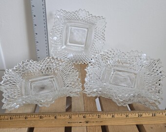 May include: Three clear, square glass dishes with a diamond pattern and scalloped edges. The dishes are arranged on a wooden surface with a ruler in the foreground. The dishes are likely for serving snacks or small portions.