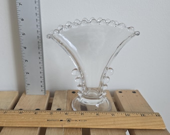 May include: A clear glass vase with a flared, fan-shaped top and a series of small, round glass beads along the rim. The vase has a round base and is approximately 6 inches tall. The vase is sitting on a wooden surface.