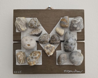 May include: A wooden plaque with a white painted letter "M" formed with various small stones. The plaque is brown and has a white painted border. The date "2024" is written in the bottom left corner and the signature "MyanSim" is written in the bottom right corner.