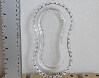 May include: Clear glass soap dish with a beaded edge. The dish is approximately 6 inches long and 3 inches wide. The dish is on a wooden surface with a metal ruler in the background.