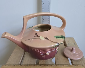 May include: A peach-colored ceramic teapot with a curved handle and spout. The teapot features a floral design with green leaves and white flowers. A matching lid sits beside the teapot. The teapot is on a wooden surface.