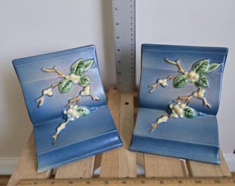 May include: Two blue ceramic bookends with a raised floral design. Each bookend features a stylized branch with green leaves and white berries. The bookends are approximately 6 inches tall and are set on a wooden surface.