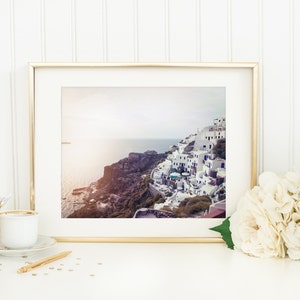 May include: A framed photograph of a white village built on a cliff overlooking the ocean. The photo is in a gold frame and is sitting on a white surface with a white hydrangea bouquet and gold coasters.