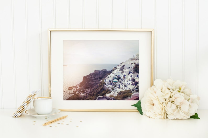 May include: A framed photograph of a white village built on a cliff overlooking the ocean. The photo is in a gold frame and is sitting on a white surface with a white hydrangea bouquet and gold coasters.