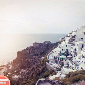 May include: A white, cliffside village with blue accents overlooking a body of water. The village is built into the side of a rocky cliff. The sky is a light blue and the water is a deep blue.