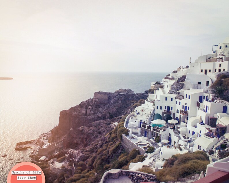 May include: A white, cliffside village with blue accents overlooking a body of water. The village is built into the side of a rocky cliff. The sky is a light blue and the water is a deep blue.