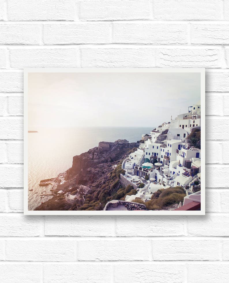 May include: A panoramic view of a whitewashed village perched on a cliff overlooking the ocean. The buildings are clustered together, with narrow streets winding between them. The sky is a soft blue, and the water is a deep turquoise.