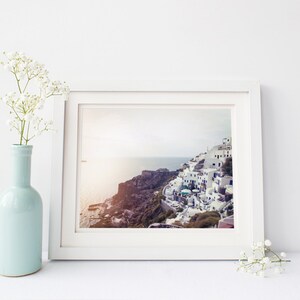 May include: A white picture frame with a photograph of a white village on a cliff overlooking the ocean. The frame is on a white surface with a vase of white flowers on the left and a sprig of white flowers on the right.