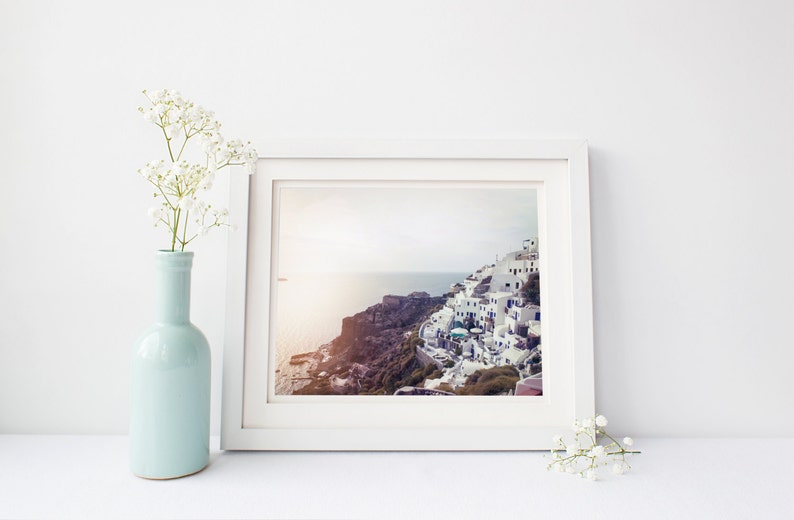 May include: A white picture frame with a photograph of a white village on a cliff overlooking the ocean. The frame is on a white surface with a vase of white flowers on the left and a sprig of white flowers on the right.