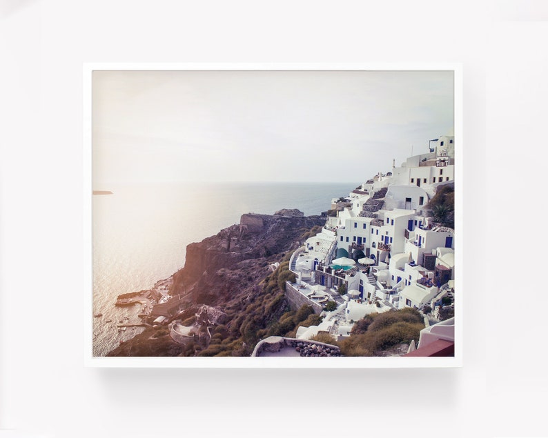 May include: A panoramic view of a whitewashed village perched on a cliff overlooking the Aegean Sea. The buildings are clustered together, with blue accents on the windows and doors. The sea is a deep blue, and the sky is a soft blue with a hint of pink.