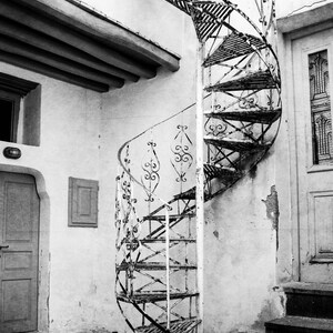 May include: A black and white photo of a weathered, metal spiral staircase leading up to a doorway. The staircase is attached to a white wall with peeling paint.