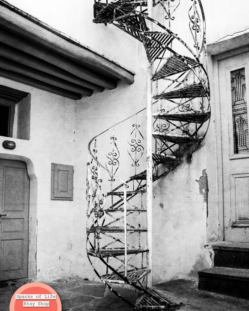 May include: A black and white photo of a weathered, metal spiral staircase leading up to a doorway. The staircase is attached to a white wall with peeling paint.