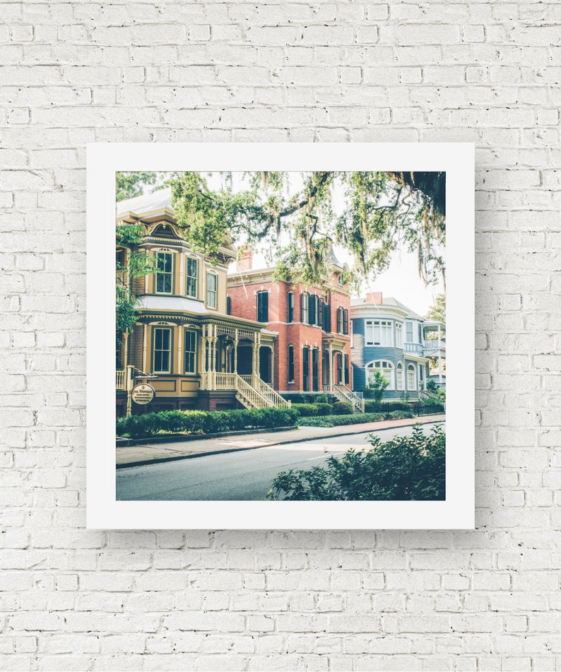 May include: A row of colorful historic houses with white trim and green foliage in front. The houses have a variety of architectural styles, including Victorian and Colonial. The houses are located on a street with a paved road and sidewalks.