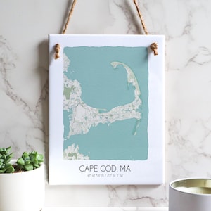 May include: A white canvas print with a light blue map of Cape Cod, Massachusetts. The map shows the outline of the Cape with roads and towns. The text "CAPE COD, MA" and "41 47 56" N / 70 16 17" W" is printed below the map.