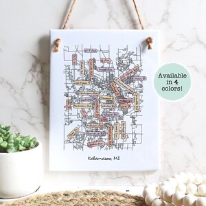 May include: A black and white line drawing of a map of Kalamazoo, Michigan. The map includes street names and landmarks. The map is printed on a white background and framed with a brown rope. The text "Available in 4 colors!" is printed in a green circle on the right side of the image. The text "Kalamazoo, MI" is printed at the bottom of the image.