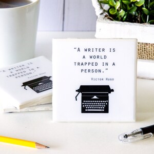 May include: White ceramic coaster with a black typewriter graphic and the quote "A writer is a world trapped in a person." by Victor Hugo.