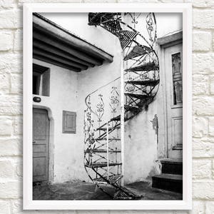 May include: A black and white photograph of a spiral staircase in a building with a white wall and a door. The staircase is made of metal and has a decorative design.