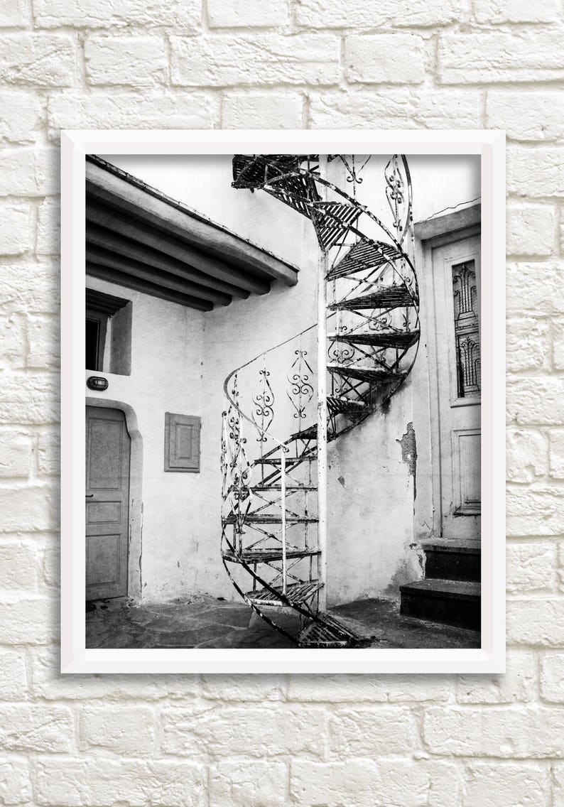 May include: A black and white photograph of a spiral staircase in a building with a white wall and a door. The staircase is made of metal and has a decorative design.
