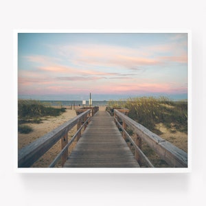 May include: A wooden walkway leads to the beach with a view of the ocean and a pink and blue sky. The walkway is lined with tall grass on either side.