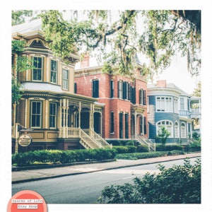 May include: A row of historic houses with colorful facades, including a yellow house with a porch, a red brick house with a white trim, and a blue house with white trim. The houses are lined up on a street with green trees and bushes in front.