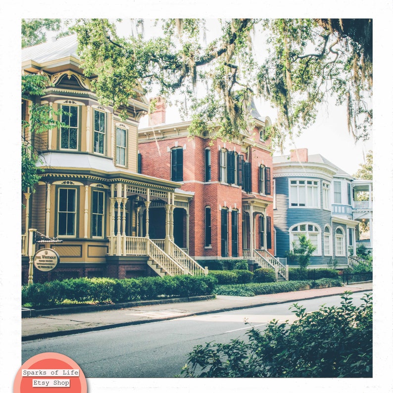 May include: A row of historic houses with colorful facades, including a yellow house with a porch, a red brick house with a white trim, and a blue house with white trim. The houses are lined up on a street with green trees and bushes in front.