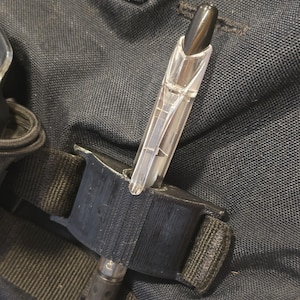 May include: A clear plastic pen is held in a black nylon holder. The pen has a black tip and a textured grip. The holder is attached to a black strap, possibly for carrying or securing the pen.
