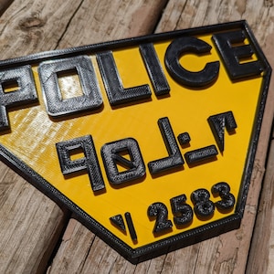 Op de afbeelding: Een geel en zwart 3D-geprint bord met het woord "POLICE" in zwarte letters en het nummer "2583" in zwarte letters.