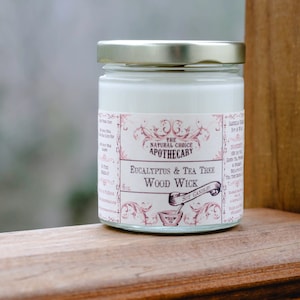 May include: A glass jar candle with a white label that reads "The Natural Choice Apothecary Eucalyptus & Tea Tree Wood Wick". The jar is sitting on a wooden surface.