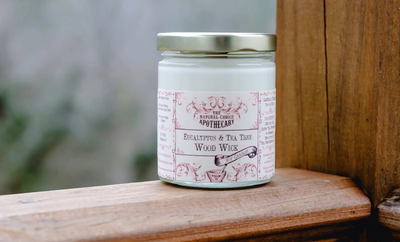 May include: A glass jar candle with a white label that reads "The Natural Choice Apothecary Eucalyptus & Tea Tree Wood Wick". The jar is sitting on a wooden surface.