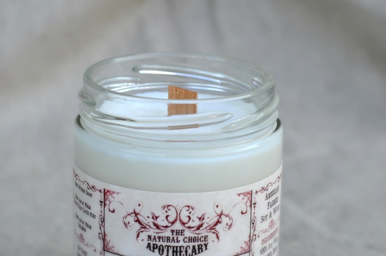 Puede incluir: Una vela blanca en un tarro de vidrio transparente con una mecha de madera. El tarro tiene una etiqueta que dice "The Natural Choice Apothecary".