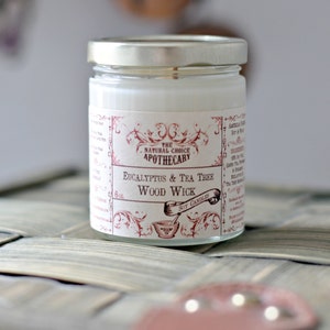 May include: A white candle in a glass jar with a red and white label that reads "The Natural Choice Apothecary Eucalyptus & Tea Tree Wood Wick Soy Candles".