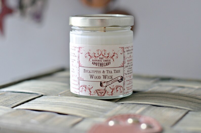 May include: A white candle in a glass jar with a red and white label that reads "The Natural Choice Apothecary Eucalyptus & Tea Tree Wood Wick Soy Candles".