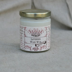 Puede incluir: Una vela de vidrio en un frasco con tapa dorada y una etiqueta blanca. La etiqueta tiene un borde decorativo y el texto "The Natural Choice Apothecary" y "Lavender Wood Wick".