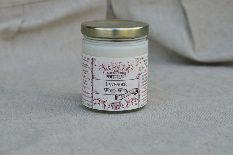 Puede incluir: Una vela de vidrio en un frasco con tapa dorada y una etiqueta blanca. La etiqueta tiene un borde decorativo y el texto "The Natural Choice Apothecary" y "Lavender Wood Wick".