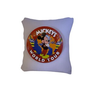 May include: A white square pillow with a large, round button featuring Mickey Mouse. The button is red with the words "Mickey's World Tour" and an image of Mickey in front of a globe.