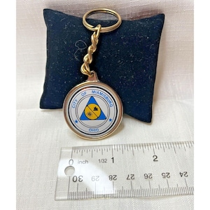 May include: Gold keyring with a white circular charm featuring the text "City of Miamisburg Ohio" and a blue triangle with a yellow sun and a white house inside. The charm is approximately 3.8 cm in diameter.