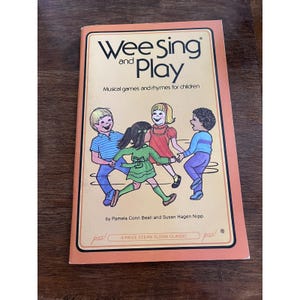 May include: A vintage children's book titled "Wee Sing and Play." The cover features a colorful illustration of four children holding hands. The book is orange with black and white text, and the title is in a stylized font. The book is a Price Stern Sloan Classic.