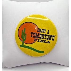 May include: A round, yellow button with the text "ENJOY A TOMBSTONE PIZZA". The button features a cactus graphic inside an orange circle, with a green line below. The button is set against a white background.