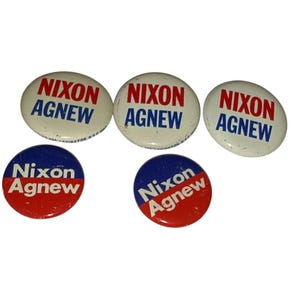 May include: A selection of vintage Nixon Agnew campaign badges. The badges are white, red, and blue, featuring the words "Nixon Agnew". The badges are in oval and circular shapes, showing signs of age.