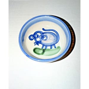 May include: A small, round ceramic bowl with a blue rim and a whimsical blue mouse illustration in the center. The mouse is painted in a simple, cartoonish style, with a green shape below it. The bowl is on a white surface.