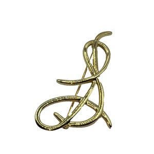 Puede incluir: Broche de metal dorado con un diseño abstracto. El broche presenta una serie de líneas curvas y en bucle que se cruzan y se superponen. El alfiler es de color dorado brillante y tiene un cierre de alfiler clásico en la parte posterior.