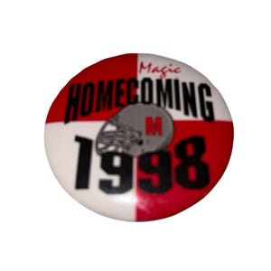 Pode incluir: Um botão redondo com um padrão xadrez vermelho e branco. O botão apresenta as palavras "Magic Homecoming 1998" em preto e um capacete de futebol cinza com um "M" vermelho no centro.